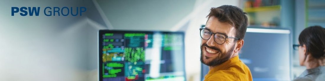 Lächelnder Mann mit Brille an einem Arbeitsplatz mit mehreren Monitoren, im Hintergrund unscharf eine weitere Person.